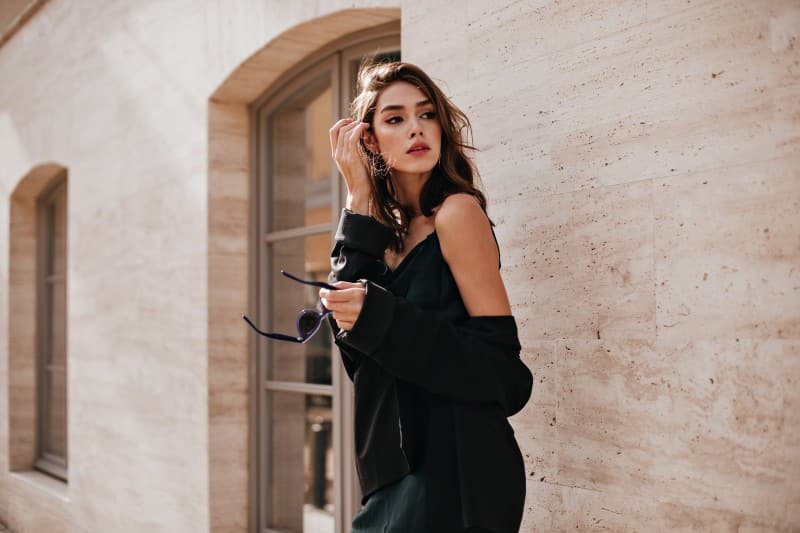 cute-young-girl-with-dark-wavy-hairstyle-bright-makeup-silk-dress-black-jacket-holding-sunglasses-hands-looking-away-against-beige-building-wall-1 Home Stylish